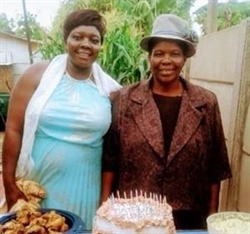 [ai] Two women stand beside a decorated cake on a table outdoors. One woman is wearing a light blue dress with a white shawl, and the other is dressed in a dark coat and hat. A plate of food is visible on the table.